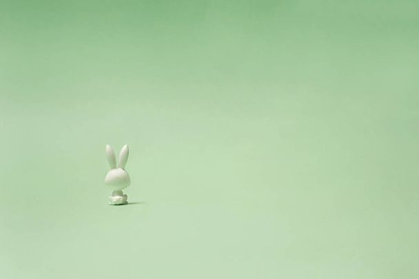 Ένα μοναχικό κουνέλι παιχνίδι κάθεται και κοιτάζει σε παστέλ πράσινη απόσταση. Φωτογραφία από την πλάτη του κουνελιού. - Φωτογραφία, εικόνα