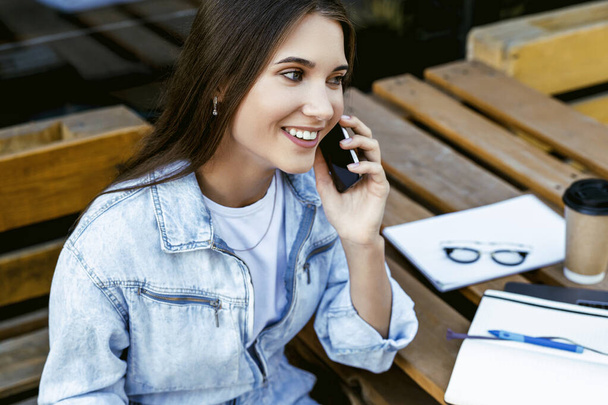 Lächelnde junge Geschäftsfrau, die in einem Café auf der Straße sitzt und mit dem Smartphone spricht. Millennium-Frau arbeitet - Foto, Bild