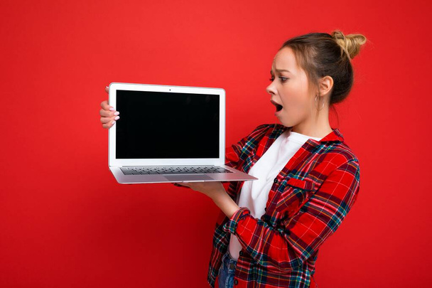 photo of Beautiful shocked amazed young woman holding laptop wearing red shirt looking at monitor display isolated on red background - Zdjęcie, obraz