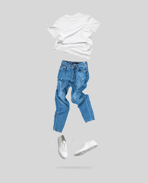 Weißes fliegendes Baumwoll-T-Shirt, blaue Jeans, weiße Lederturnschuhe auf grauem Hintergrund. Sauberes weißes Unisex T-Shirt. Markenkleidung. Mock up für Ihr Design. Frühling Sommer Damenbekleidung. - Foto, Bild