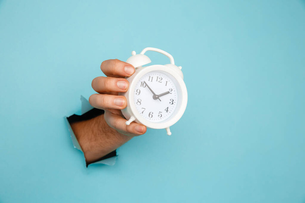 Hand holds alarm clock through a paper hole. Time management and deadline concept - Fotó, kép