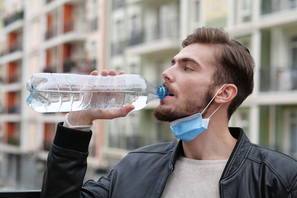 Молодой красивый мужчина в медицинской защитной маске на подбородке пьет минеральную воду из большой синей пластиковой бутылки - Фото, изображение