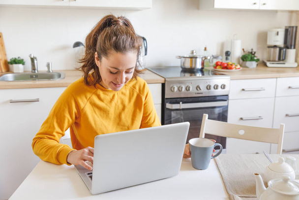 Oficina Móvil en casa. Mujer joven sentada en la cocina en casa trabajando con un ordenador portátil. Chica de estilo de vida estudiando o trabajando en interiores. Concepto de cuarentena empresarial independiente - Foto, imagen