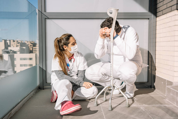 Uitgeputte mannelijke en vrouwelijke artsen in maskers en uniform zittend op de vloer op het balkon van de kliniek en pauzes nemend tijdens de harde werkdag tijdens het coronavirus - Foto, afbeelding