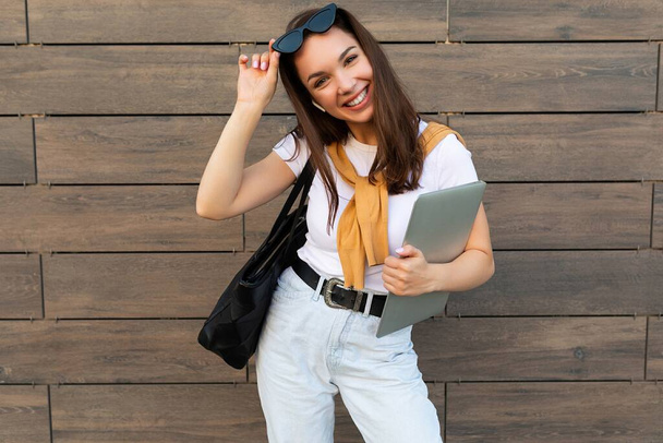 Foto der schönen Frau trägt weißes T-Shirt mit Computer-Laptop und schwarzer Sonnenbrille, die auf der Straße bleibt und in die Kamera schaut - Foto, Bild