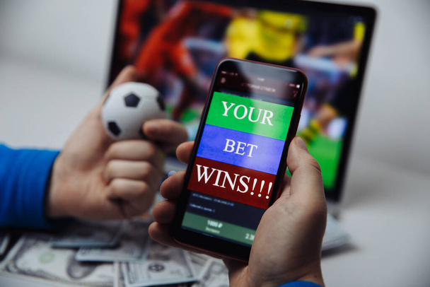 Mann sieht Fußballspiel online Übertragung auf seinem Laptop und hält Smartphone mit Online-Glücksspiel-Anwendung und Gewinnnachricht. Smartphone im Fokus - Foto, Bild