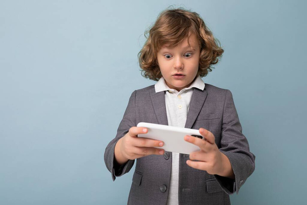 Photo shot of Handsome emotional child boy with curly hair wearing grey suit holding and using phone isolated over blue background looking at smartphone playing games - Photo, Image