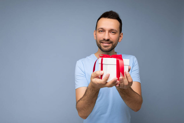 Photo shot of handsome positive brunette unshaven young man isolated over blue background wall wearing blue t-shirt holding white gift box with red ribbon and looking at camera - Photo, Image