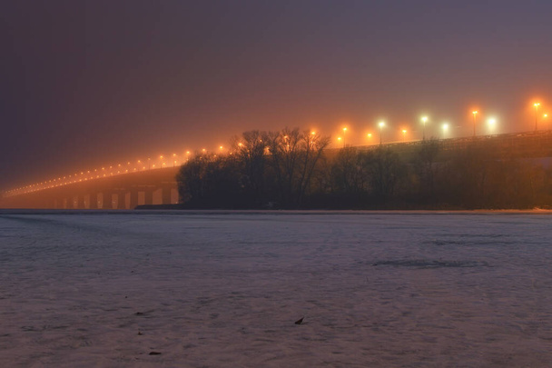 Misterioso paisaje invernal del famoso puente Paton sobre el congelado río Dnipro. Puente de iluminación de luz nocturna en una noche de niebla. Kiev, Ucrania. - Foto, imagen