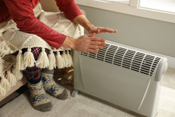Man warming hands near electric heater at home, closeup - Фото, изображение