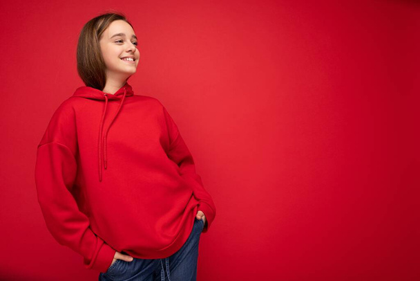 Фотография привлекательной счастливой улыбающейся брюнетки девушки-подростка в стильной красной толстовке, стоящей изолированно на красном фоне, смотрящей в сторону - Фото, изображение