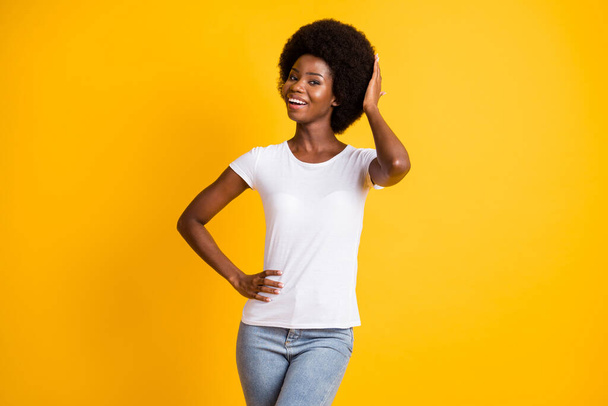 Photo portrait of young woman touching her afro hairstyle with one hand other on waist smiling wearing white t-shirt isolated on bright yellow colored background - Foto, imagen