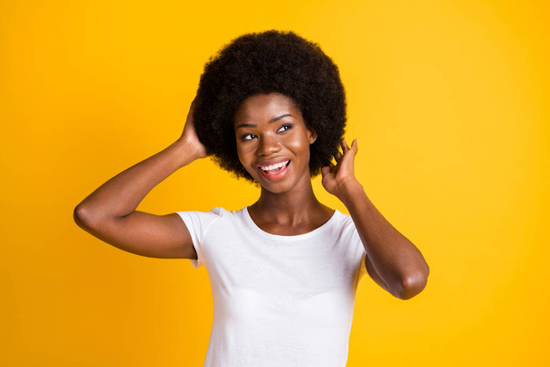 Photo portrait of young african american woman touching afro hairstyle with two hands smiling wearing white t-shirt isolated on vivid yellow colored background - Foto, Imagem