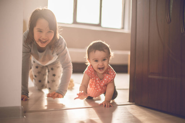 Κορίτσι παίζει με τη μικρή αδελφή της στο σπίτι. Διασκεδάζουν όταν περπατάνε στα τέσσερα. - Φωτογραφία, εικόνα