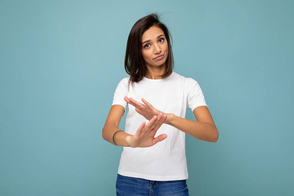 Portret młodej pięknej brunetki ze szczerymi emocjami ubranej w modny biały t-shirt na makietę odizolowany na niebieskim tle z pustą przestrzenią i pokazujący gest timeouta - Zdjęcie, obraz