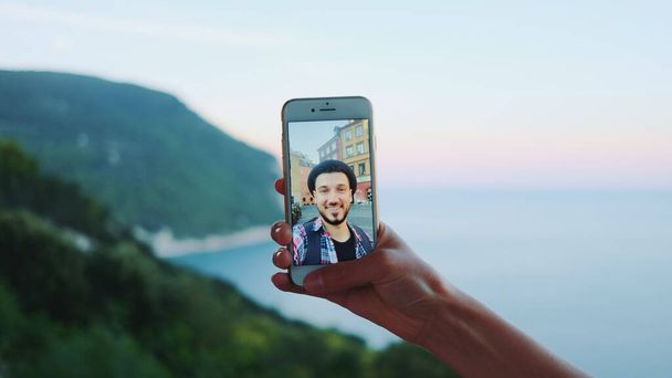Κοντινό πλάνο του hand holding smartphone κατά τη διάρκεια βιντεοκλήσης με τον άνθρωπο μπροστά στη θάλασσα - Φωτογραφία, εικόνα