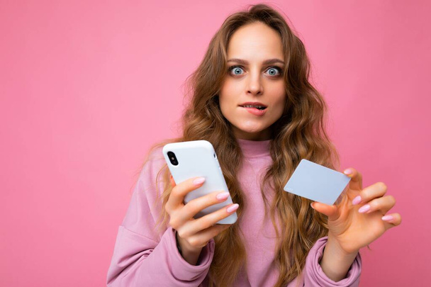 Closeup portrait photo of pretty shocked young blonde curly woman wearing pink clothes isolated over pink background using mobile phone making payment using credit card looking at camera and having - Photo, Image