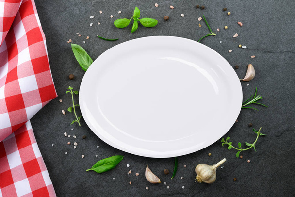 Klasická italská kuchyňská šablona - horní pohled na bílý prázdný keramický talíř na vinobraní černého kamene kuchyňský pult s červeným kostkovaným ubrusem a výběr bylin a koření. - Fotografie, Obrázek