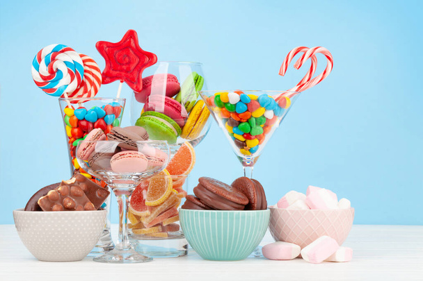 Różne słodycze asortyment. Cukierki, bonbony, czekolada i makaroniki w kieliszkach koktajlowych na niebieskim tle - Zdjęcie, obraz