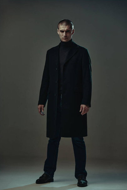 Confident serious angry guy in black coat, detective or security on gray studio background, full-length male portrait - Φωτογραφία, εικόνα