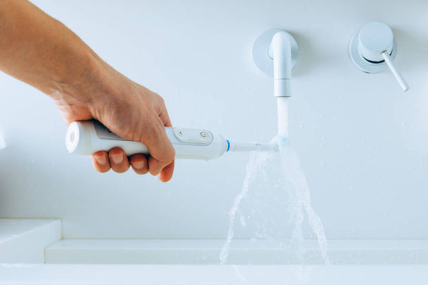 mano sosteniendo un cepillo de dientes bajo el agua que fluye del grifo en un baño - Foto, imagen