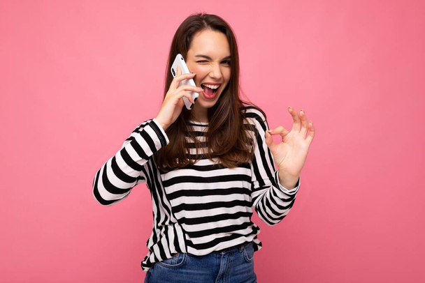 Vinkkaaminen Melko onnellinen nuori nainen puhuu puhelimessa yllään raidallinen villapaita eristetty taustan yli kopioi tilaa osoittaa ok ele katsot kameraa - Valokuva, kuva
