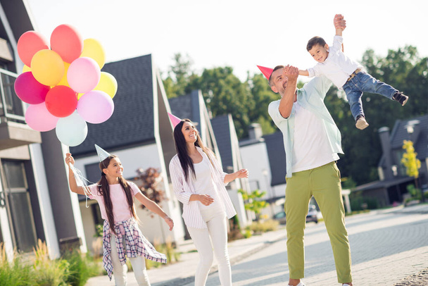 Photo portrait of married couple with children wearing party cones. Father throws boy up in air outdoors - Фото, изображение