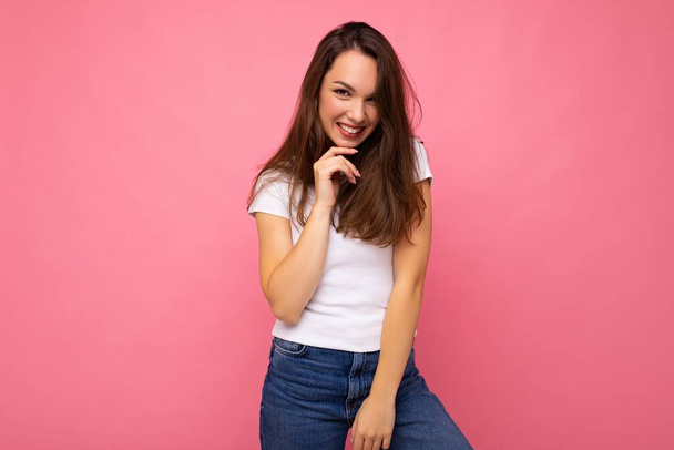 Photo portrait of young beautiful smiling hipster brunette woman in white t-shirt with mockup. Sexy carefree female person posing isolated near pink wall with empty space in studio. Positive model - 写真・画像