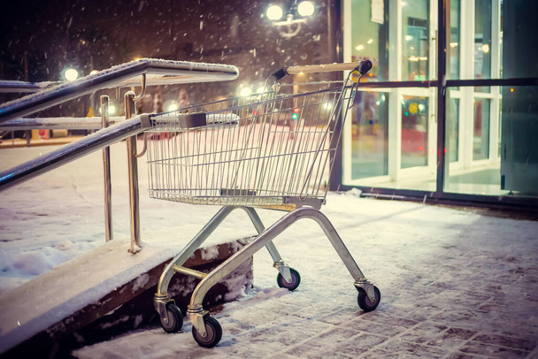 Leeg winkelwagentje staat op de trappen buiten de winkel op een besneeuwde winteravond - Foto, afbeelding