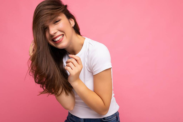 Young beautiful brunet woman. Trendy lady in casual summer white t-shirt for mock up. Positive female shows facial sincere emotions. Funny model isolated on pink background with free space - 写真・画像