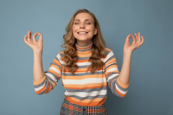 Portrait of smiling happy young attractive blonde curly woman with sincere emotions wearing trendy striped pullover isolated on blue background with copy space and doing mediation gesture with fingers - Fotografie, Obrázek