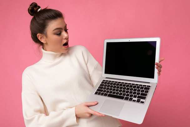 Photo of beautiful amazed and surprised woman with gathered brunette hair wearing white sweater holding computer laptop and looking at the open netbook isolated over pink wall background - Foto, Imagen
