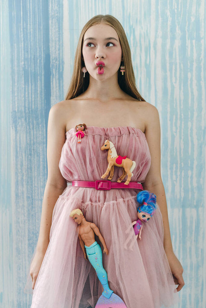 Portrait of young girl wearing pink dress and belt with dolls on blue wall background. Barbie style, posing concept  - Zdjęcie, obraz