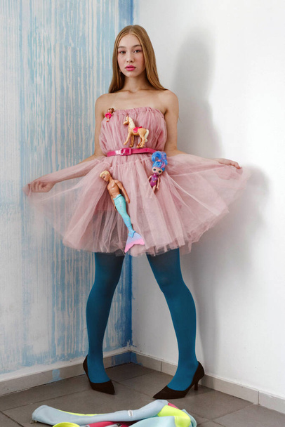 Girl model in pink dress with dolls on blue and white wall background. Barbie style, posing concept - Foto, Imagen