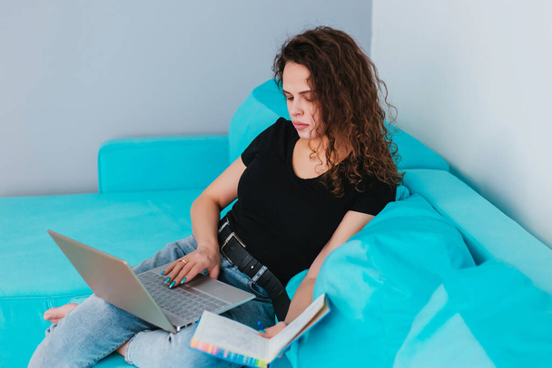 Egy fiatal és gyönyörű nő laptopot használ, szöveget ír, egy kék kanapén ül, szövegíróként dolgozik. Keresi a számítógépen keresztül az interneten, blogok, tanulmányok otthon - Fotó, kép