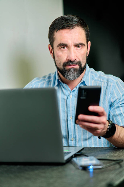 homem branco com barba e camisa azul t trabalhando com telefone celular e computador - Foto, Imagem