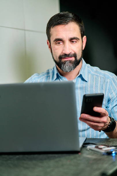 witte man met baard en blauw t-shirt werkend met mobiele telefoon en computer - Foto, afbeelding