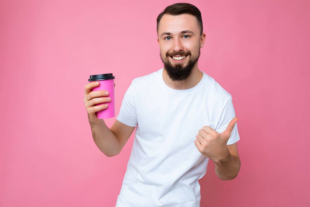 photo of handsome smiling young brunette unshaven male person with beard wearing white t-shirt isolated over pink backgroung wall holding paper coffee cup drinking and looking at camera and showing - Φωτογραφία, εικόνα