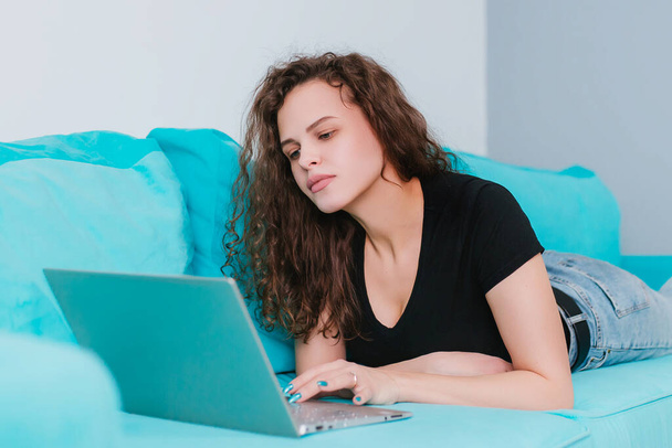 Una mujer joven y hermosa utiliza una computadora portátil, comprueba las noticias por correo electrónico mientras está acostada en un sofá azul, buscando amigos en las redes sociales. Trabaja en una computadora a través de Internet, escribe un blog, estudia en casa o ve un webinar - Foto, imagen