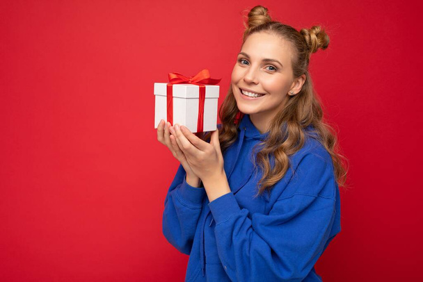 Beautiful happy young blonde woman isolated over colourful background wall wearing stylish casual clothes holding gift box and looking at camera - Zdjęcie, obraz