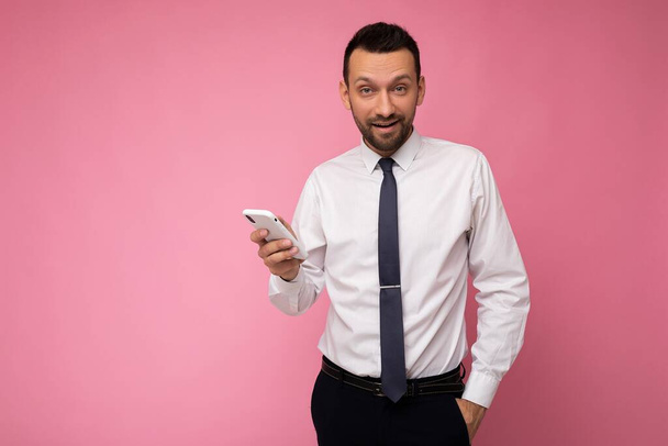 Fénykép jóképű jóképű férfi visel alkalmi fehér ing és nyakkendő elszigetelt rózsaszín háttér üres hely tartja a kezében, és a mobiltelefon üzenetküldő sms nézi a kamerát - Fotó, kép