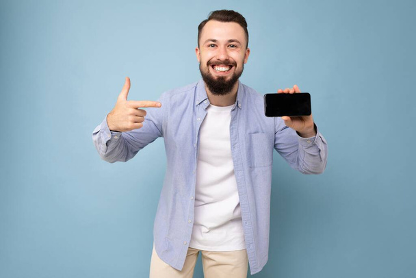 Photo of handsome smiling adult male person good looking wearing casual outfit standing isolated on background with copy space holding smartphone showing phone in hand with empty screen display for - Photo, Image