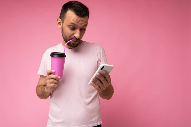 Foto von hübschen jungen unrasierten brünetten Mann mit Bart trägt jeden Tag hellrosa T-Shirt isoliert über rosa Hintergrund halten und mit dem Handy im Internet surfen und trinken - Foto, Bild