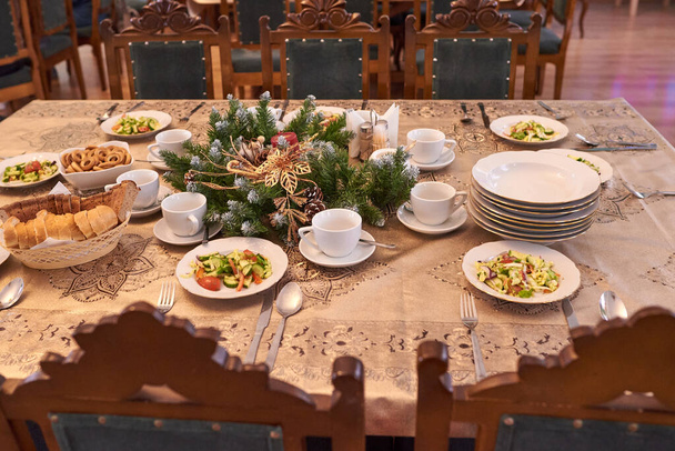 スプルースの枝やコーンで飾られた新年のテーブル。白いカップとソーサー、プレート、パンボックス、ベーグル、野菜サラダ - 写真・画像
