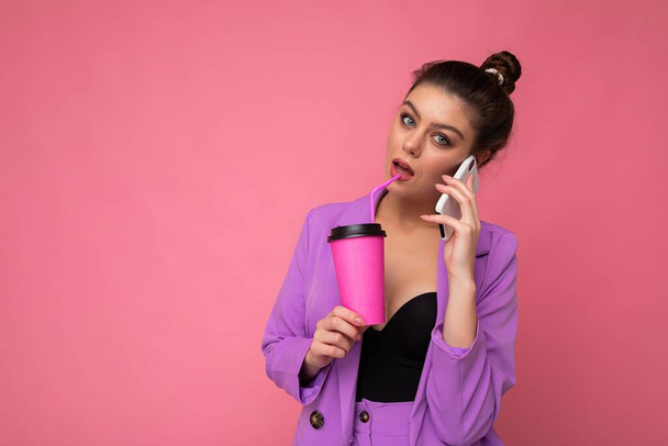 Портрет красивой брюнетки носить модный фиолетовый пиджак, говорить по смартфону, держа кофе на вынос в бумажной чашке, изолированные над розовым фоном пустое место для рекламы - Фото, изображение