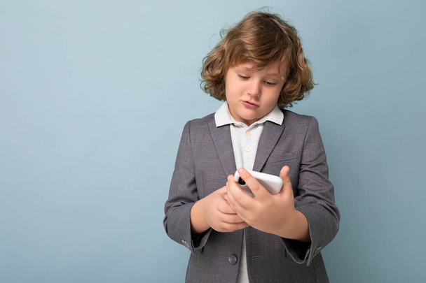 Zdjęcie Przystojnego małego chłopca z kręconymi włosami w szarym garniturze trzymającego i korzystającego z telefonu odizolowanego na niebieskim tle patrzącego na smartfona komunikującego sms-y z filmu - Zdjęcie, obraz