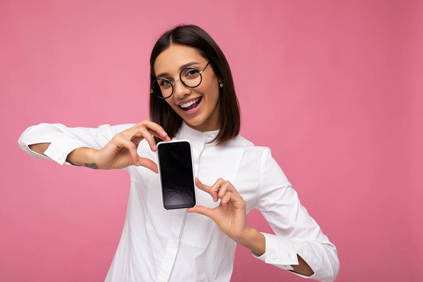 Photo of beautiful smiling young woman good looking wearing casual stylish outfit standing isolated on background with copy space holding smartphone showing phone in hand with empty screen display for - Photo, Image