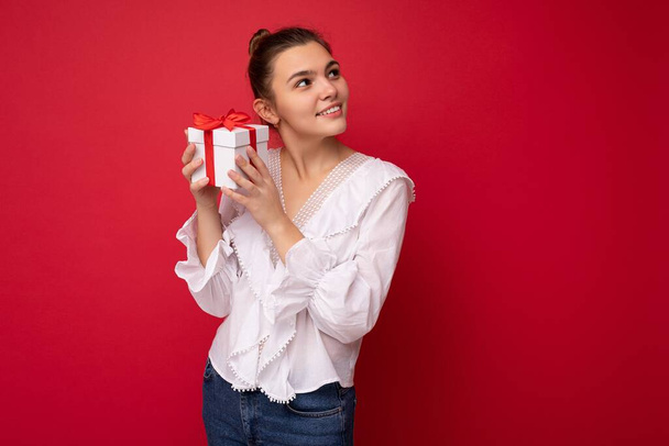 Shot of charming happy smiling young brunette woman isolated over red background wall wearing white blouse holding white gift box with red ribbon and looking to the side - Foto, afbeelding