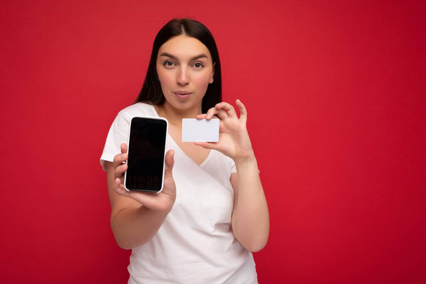 Foto van aantrekkelijke jonge brunette vrouw met casual wit t-shirt geïsoleerd over rode achtergrond met lege ruimte in de hand mobiele telefoon en toont smartphone met leeg scherm voor - Foto, afbeelding