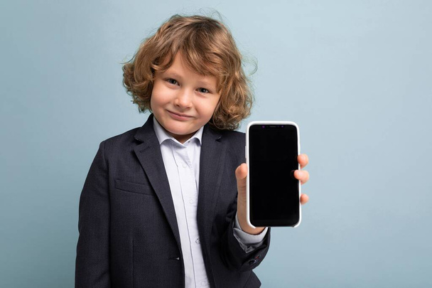Closeup photo of Handsome positive boy with curly hair wearing suit holding phone isolated over blue background looking at camera and showing smartphone with empty display screen - Photo, Image